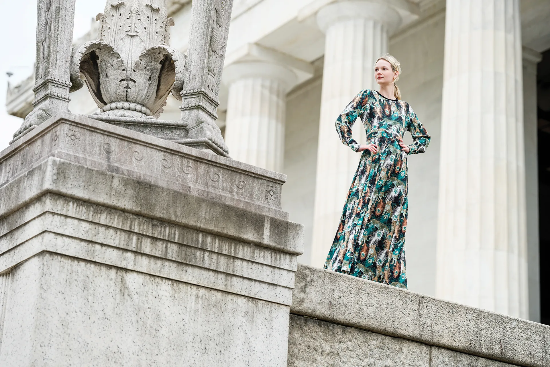 Photoshoot of blonde woman wearing a long dress with peacock feather pattern standing in front of old white building with white columns