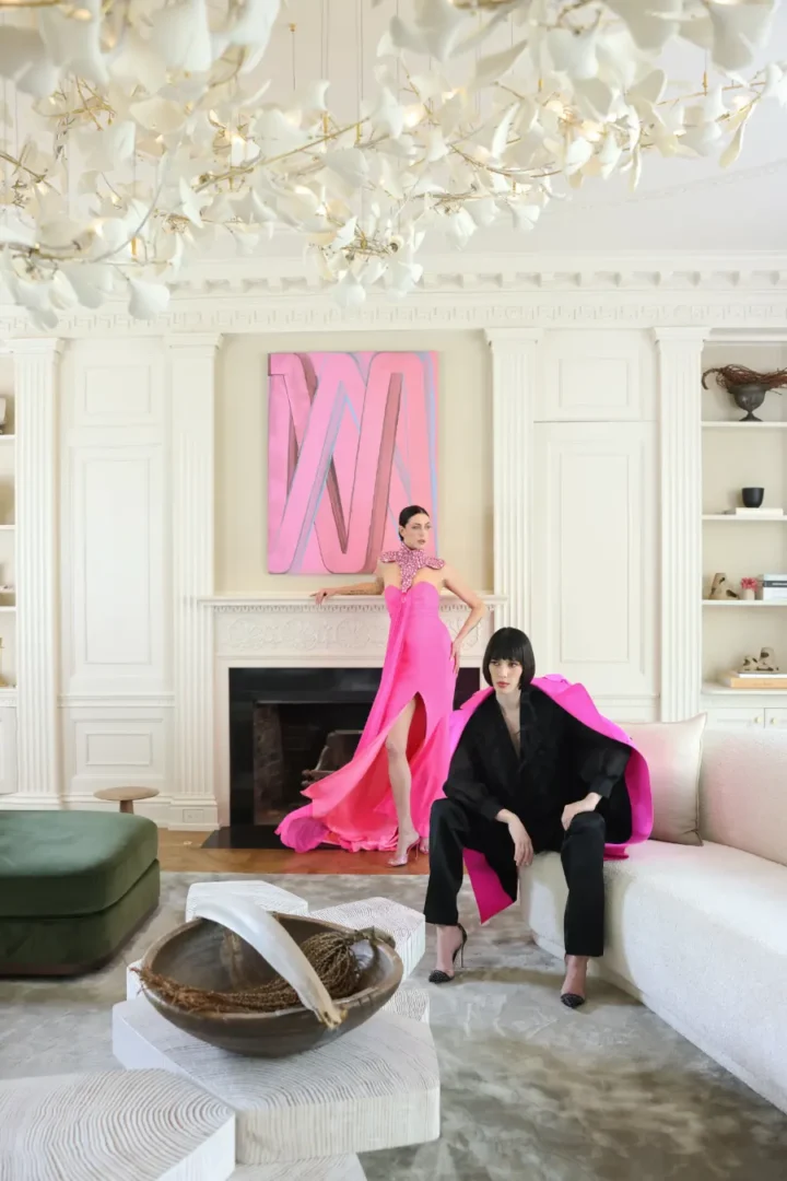 photo shoot inside a large light colored living room with two young ladies with black hair dressed for a party, one in a hot pink long dress with a slit in the middle, and other with a black pant suit and hot pink blazer