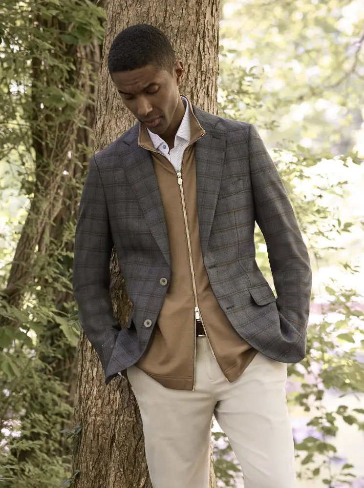 Young black man wearing a dark brown grayish blazer leaning against a tree