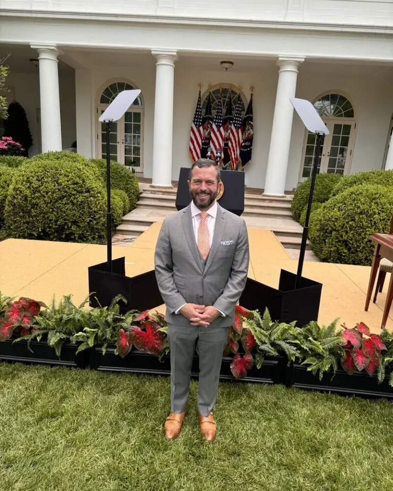 man with beard wearing gray suit with white shirt standing in grass outside of press event