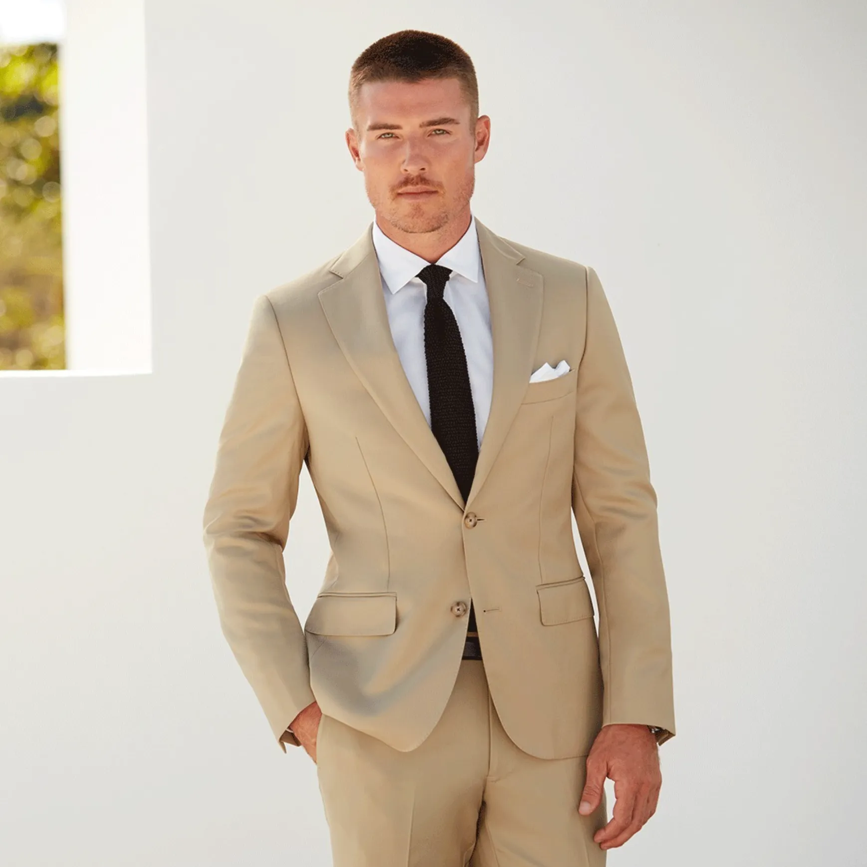 young man with buzz cut hair and blue eyes wearing a light cream-colored suite and black tie standing in front of white wall