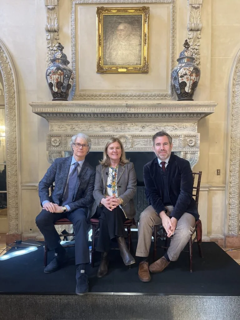 two older men dressed up wearing fall blazers and tie and woman in the middle wearing a gray blazer and black pants all sitting in chairs in front of a stone fireplace